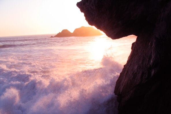 Sunset behind wave crashing onto beach rocks